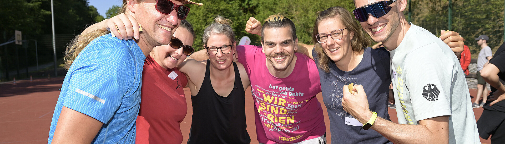 Symbolbild: Eine Gruppe von Menschen in Sportbekleidung steht lächelnd zusammen auf einem Sportplatz im Freien