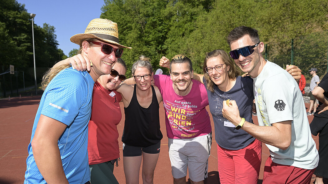 Symbolbild: Eine Gruppe von Menschen in Sportbekleidung steht lächelnd zusammen auf einem Sportplatz im Freien